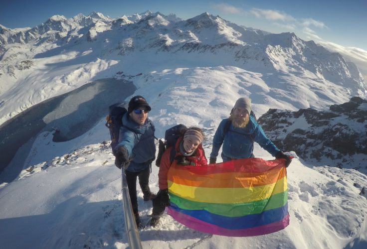 Three people on a snowy mountain top holding a rainbow flag