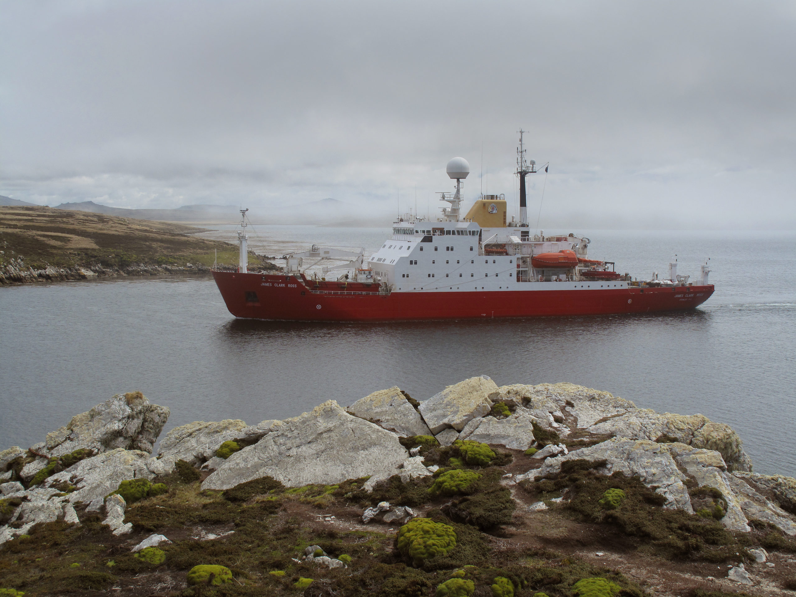 Large red ship in a body of water with rocks in the foreground