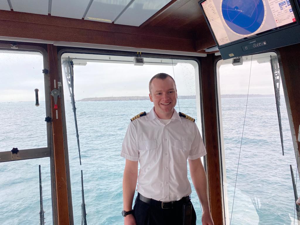 A man wearing a white shirt with black shoulder pads stands in front of a window with the sea behind him