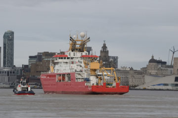 A large red ship on a body of water, with Liverpool city in the background