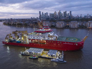 A large red ship is floating in a river with a city skyline behind it.