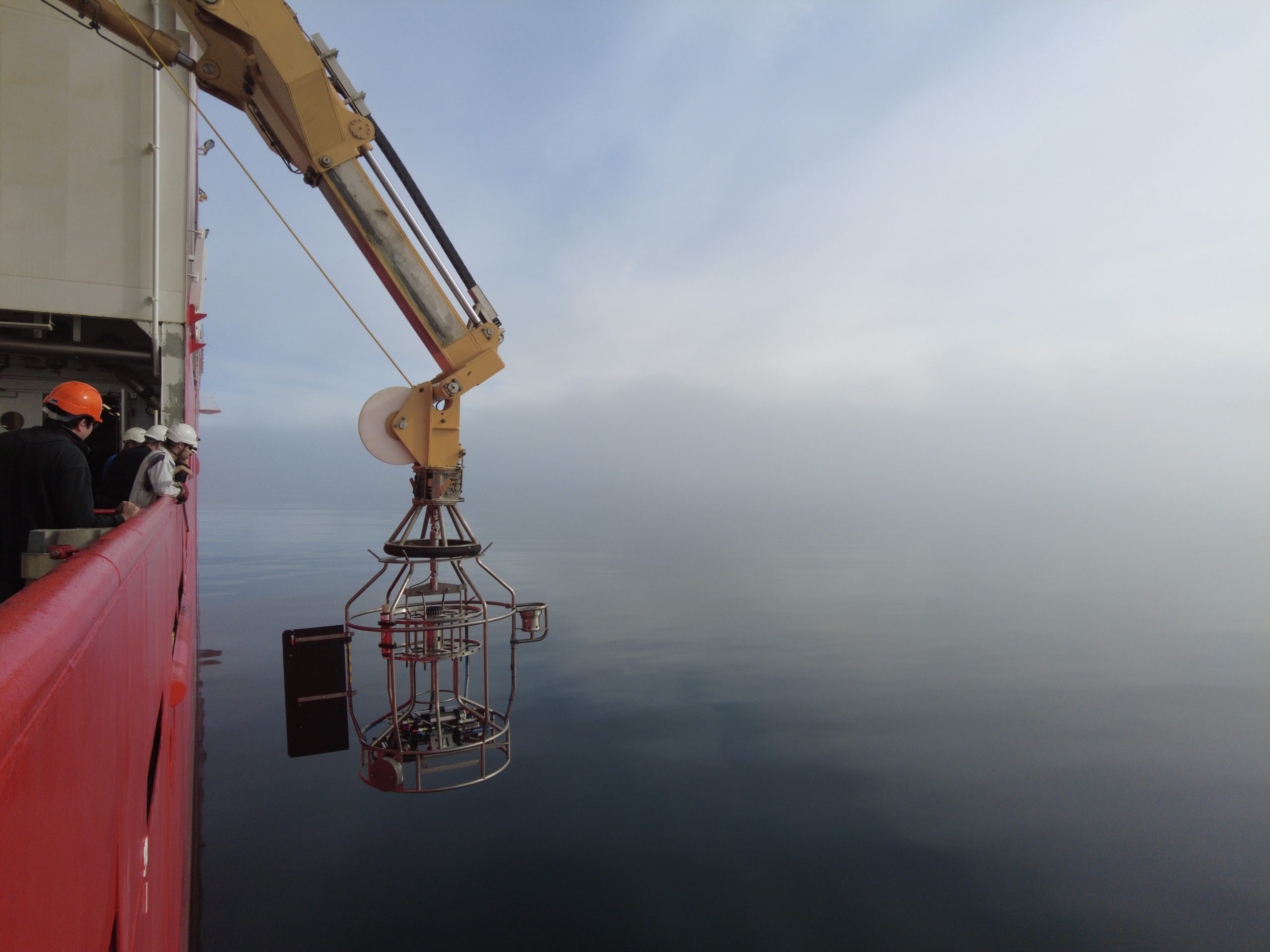 A metal scientific instrument is hanging over the side of RRS Sir David Attenborough from a yellow crane arm. The side of the ship is red, and there are people wearing hard hats looking over the side.