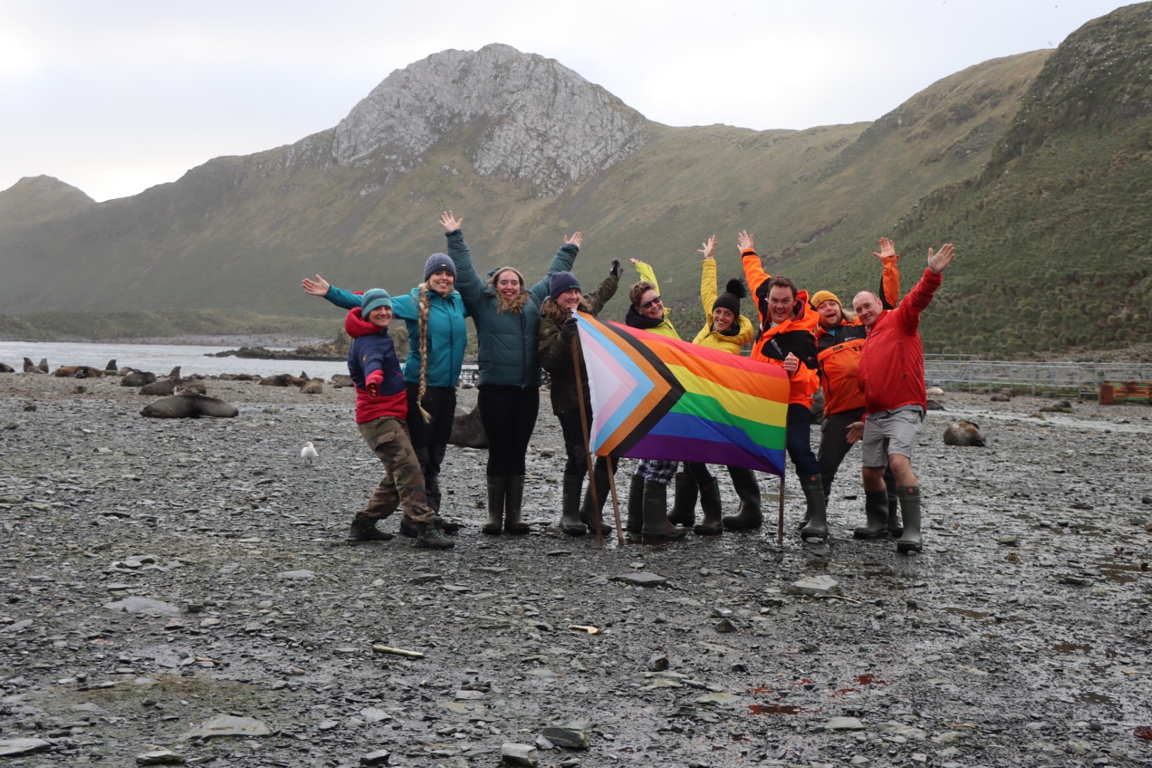 A group of 9 people stand on a stony beach holding a Pride flag. There are green peaks in the background.