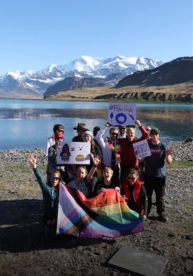 A group of 12 people are standing together holding up signs saying Polar Pride and a rainbow flag. There is water in the background, as well as green and snowy peaks.