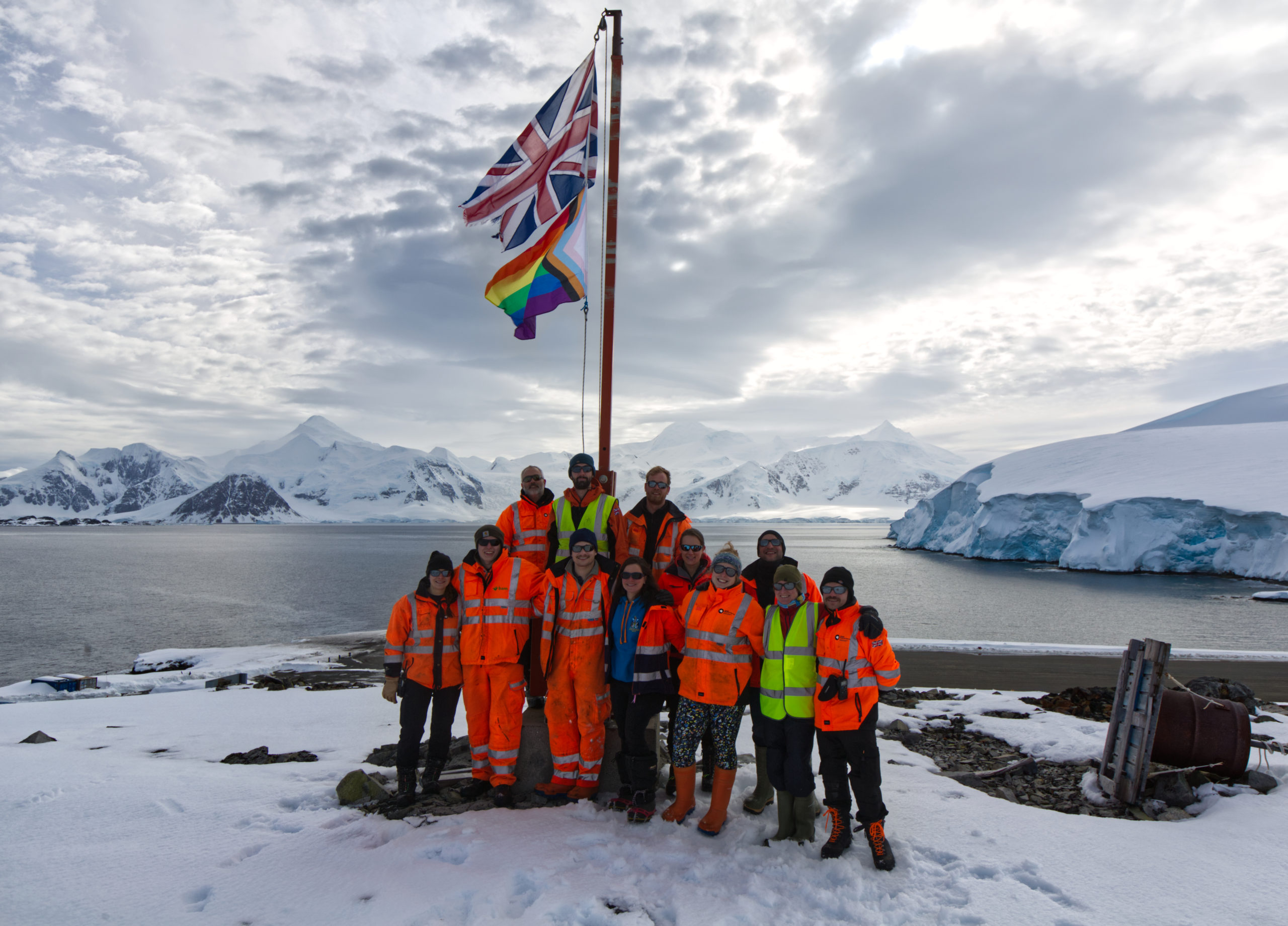 A group of 11 people wearing hi-vis coats stand on snow around a flag pole which has two flags flying from it - one is a union jack the other is a rainbow flag. In the background is a body of water and snowy mountains. 