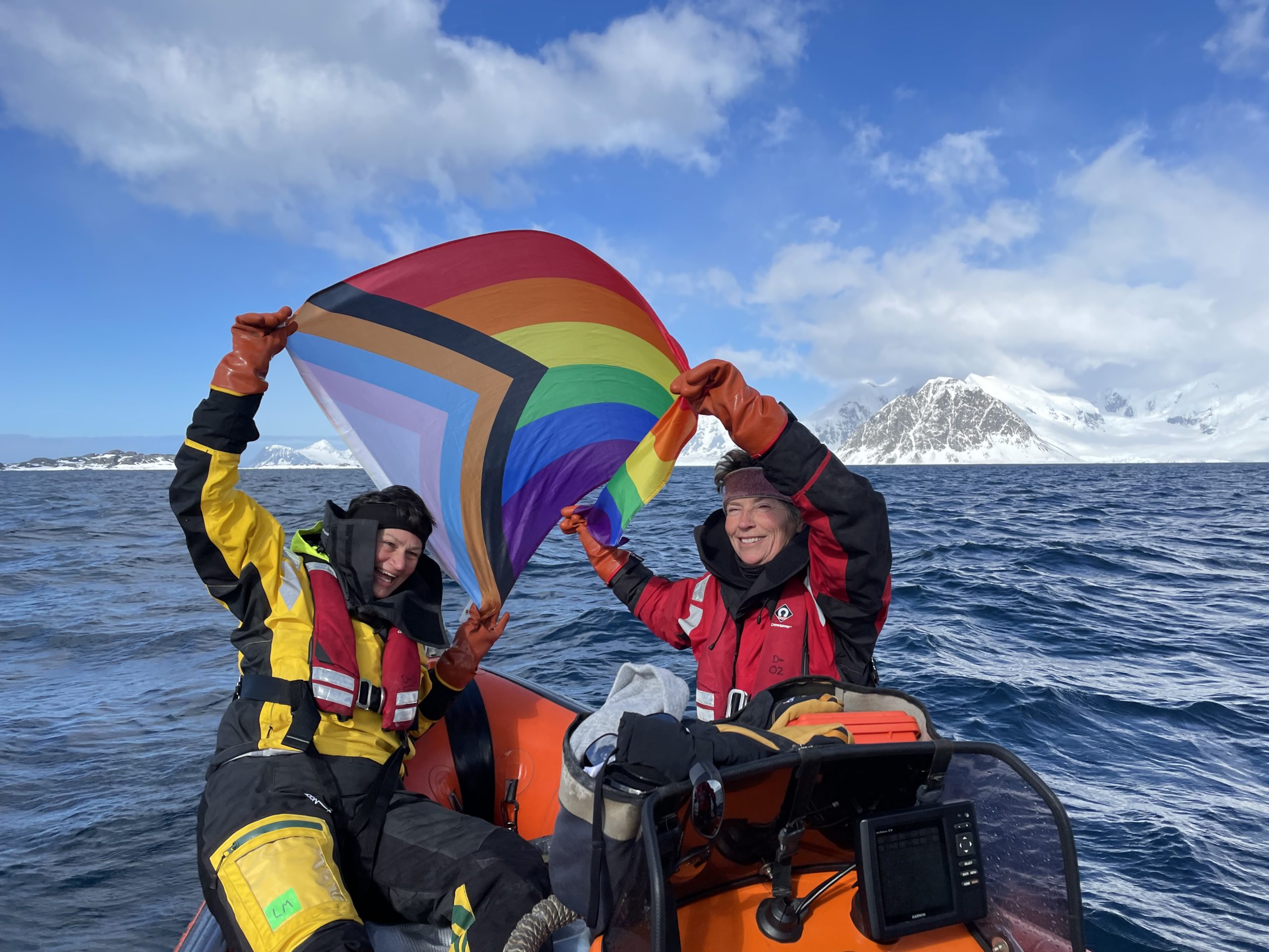 Two people are sat in a small boat holding up a rainbow Pride flag. There is a snowy mountain in the back.