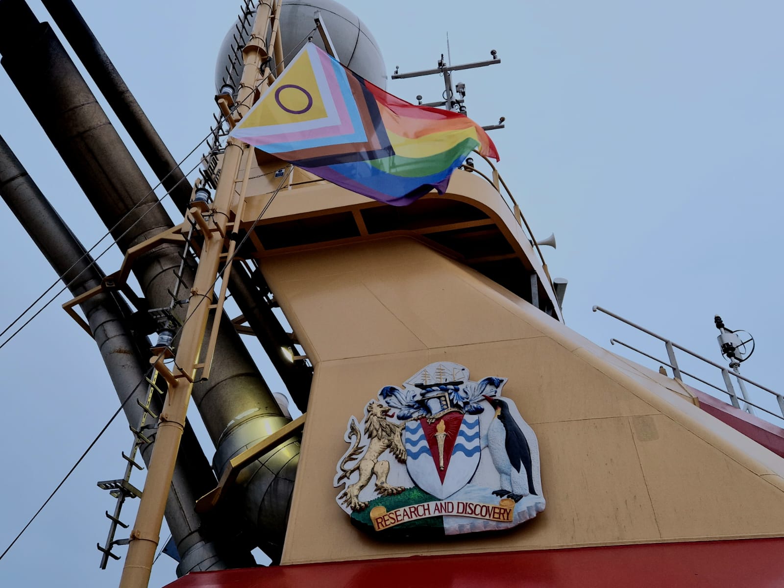 The Pride flag flies in front of a ship's superstructure.