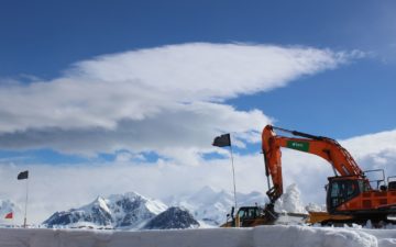 orange excavator with BAM company logo on the side clearing snow in Antarctica against a backdrop of a cloudy and blue sky with snow covered mountains in the background