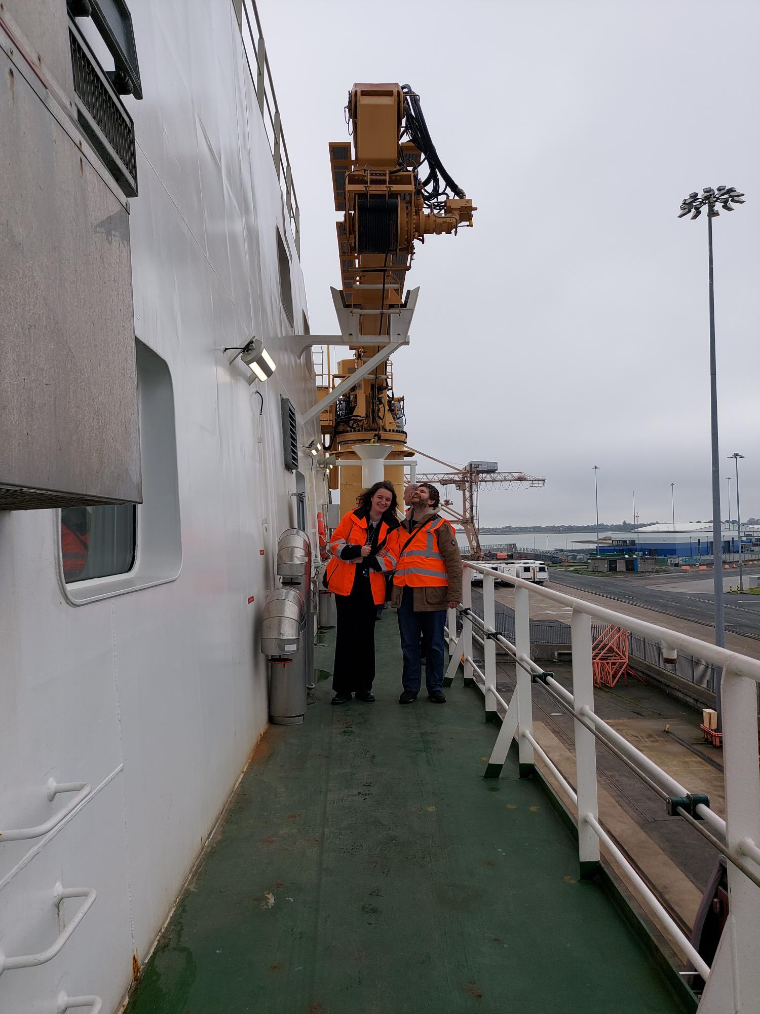 Two people wearing orange hi-vis jackets stand on the side of a ship. There is a large yellow crane behind them, the deck is green and there is a white rail to their left.