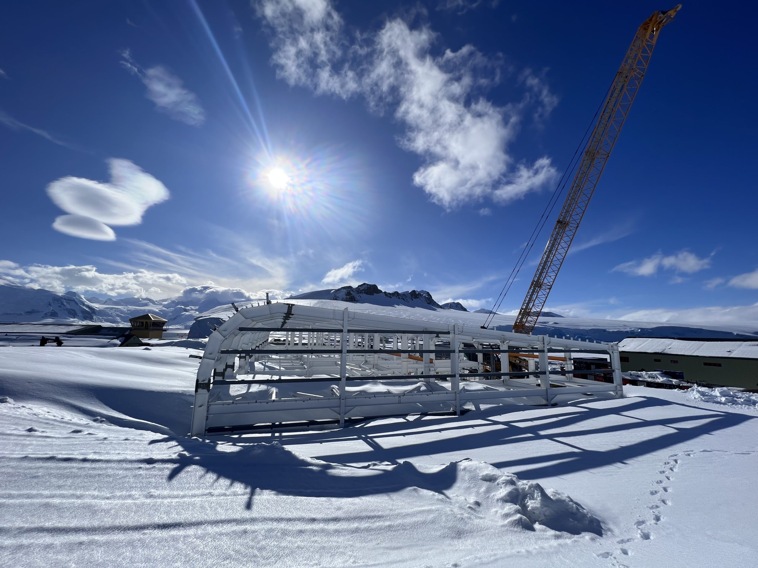 skeleton of a building being built in Antarctica with blue skies and snow in the foreground against a white steel frame with a red crane beside it