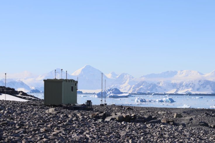 A small cube beach hut in muted colours sits against a bright blue Antarctic sky