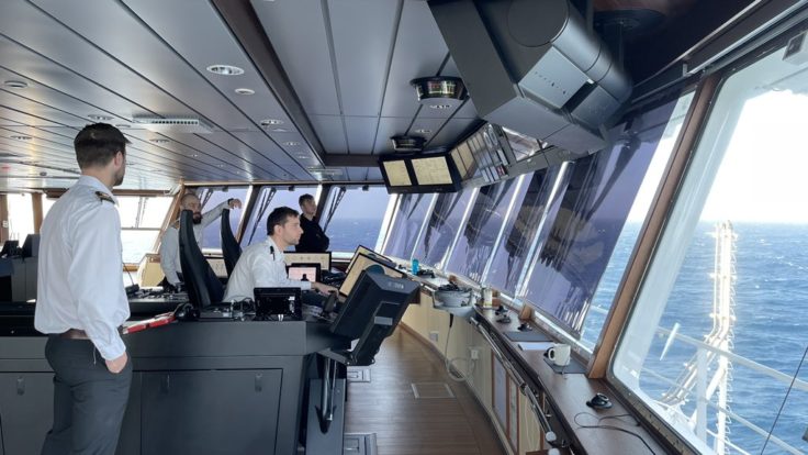 Four men stand on a large viewing platform of the research ship, one of whom is using controls to steer the vessel. Outside the windows there is a wide stretch of dark blue sea. 