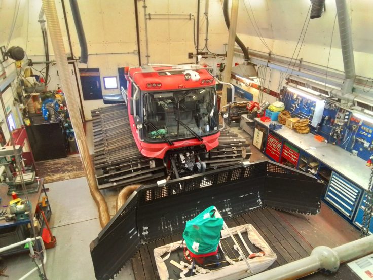 A large white garage space with a big orange plough in the middle, surrounded by work benches and tools.