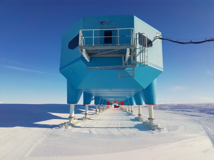 The front of a chain of blue pods stands boldly against a bight blue sky. The chain of pods is elevated on long, extended skis. The show is ploughed smooth and flat.