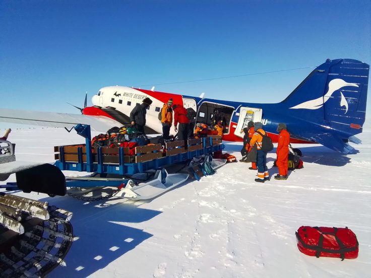 A group of 10 people dressed in orange warm outwear load a sledge of bags and equipment into a small blue and red plane.