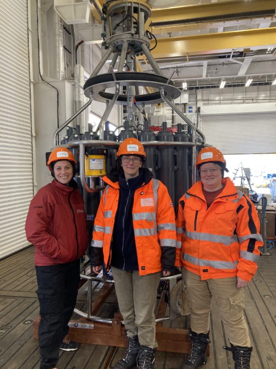 Three scientists in high visibility jackets stand in front of the large CTD instrument - a cylinder structure filled with slim sample tanks.