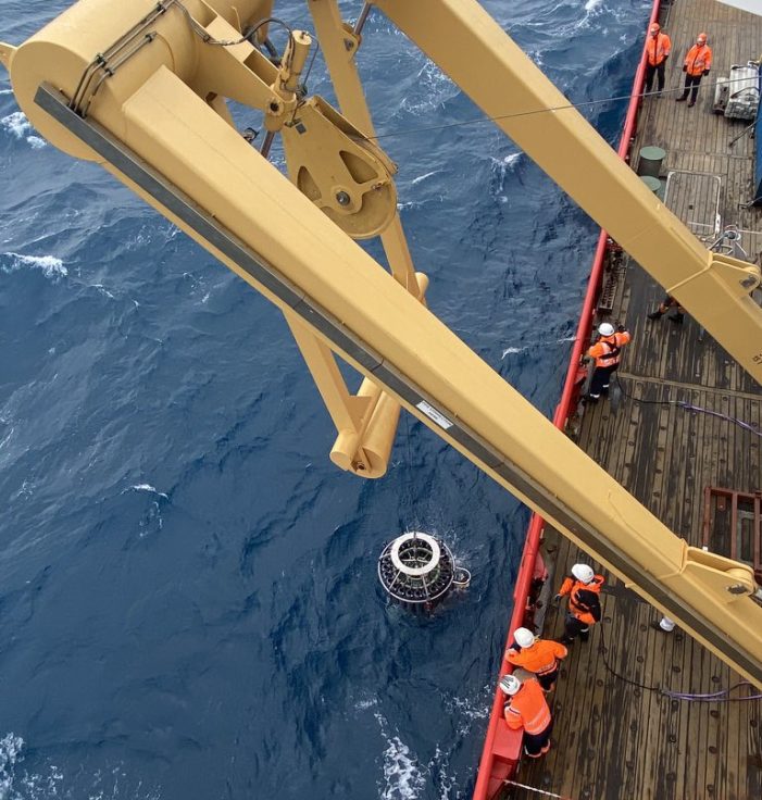 Viewed from above, members of the science team, dressed in orange, watch a CTD being lowered into the sea by a large yellow crane.