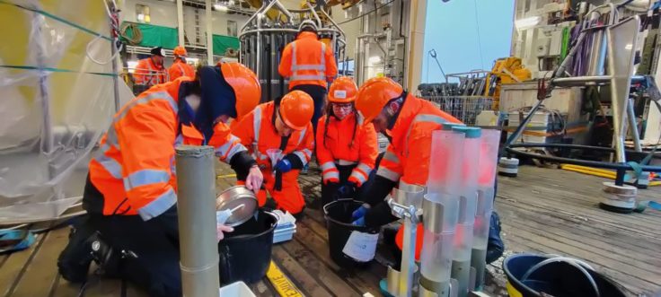 A group of scientists kneel together on the deck of the RRS Sir David Attenborough, handling sediment samples from a series of clear tubes taken from the Multicorer.
