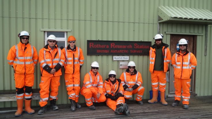 A group of people wearing orange safety visibility clothing standing outside a green building