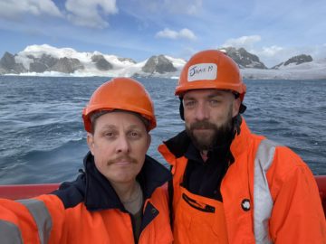 Two men dressed in orange hi vis jackets and hard hats pose for a selfie, in front of snow-covered mountains and the sea in the foreground.