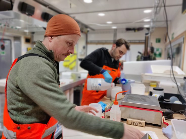 Two men work in the wet lab aboard the ship. They wear orange waterproof trousers and warm fleeces. They are sorting samples from the trawl.