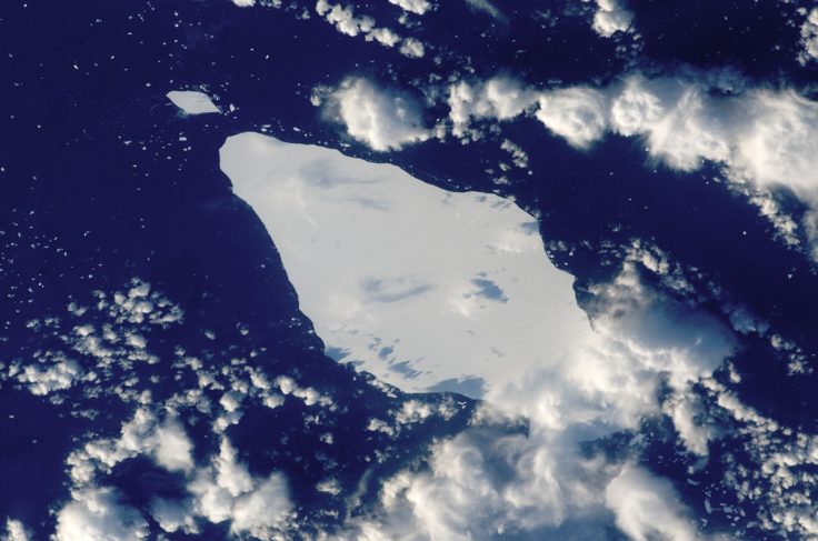 A diamond shaped iceberg in a deep blue sea, viewed from space. Clouds are dotted across the picture, obscuring the view of the Earth's surface in places.