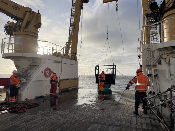 Four people in orange hi-vis lower a scientific trawling net off the deck of the ship with a large yellow crane and winch.