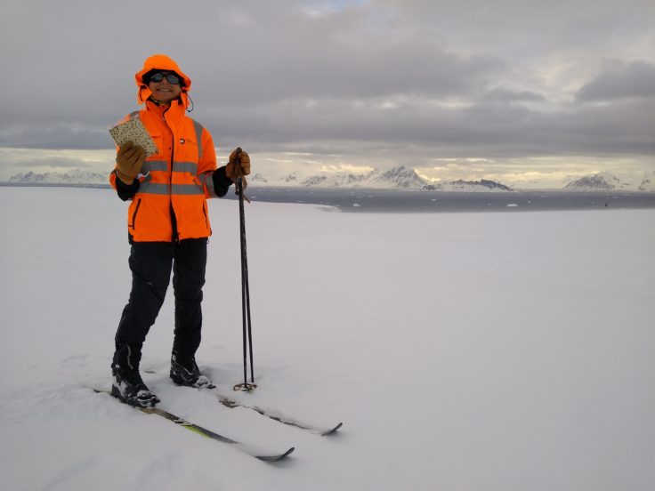 Alysa is standing in a snowy landscape wearing skis, a high visibility warm jacket and holding a large matza cracker.
