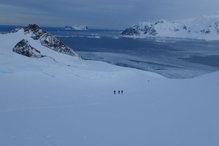 A wide, open Antarctic valley, full of snow and looking down to the sea. Three figures are visible ski treking up the hill in the distance.