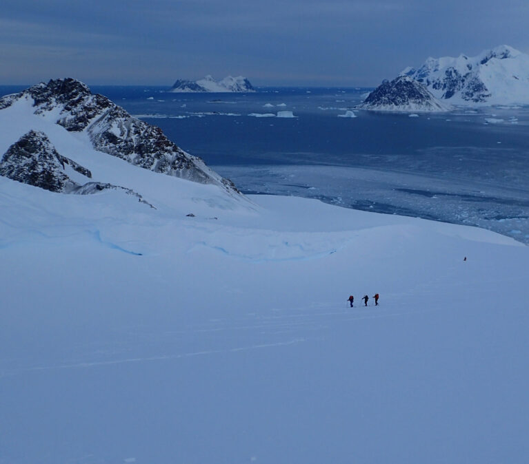 A wide, open Antarctic valley, full of snow and looking down to the sea. Three figures are visible ski treking up the hill in the distance.