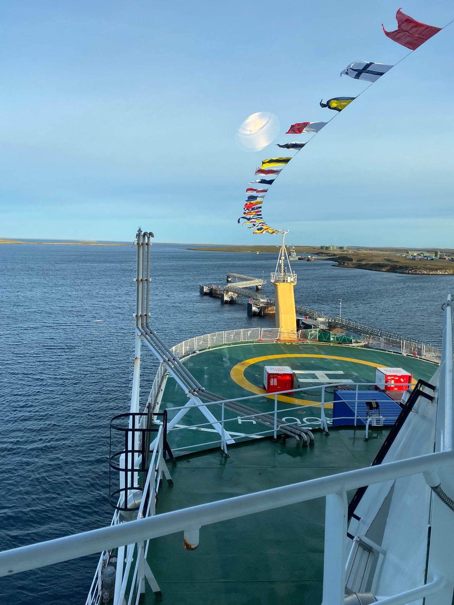 This picture looks out over the helideck of a ship. There are a line of flags with different colours and patterns that are streaming in the wind over the helideck