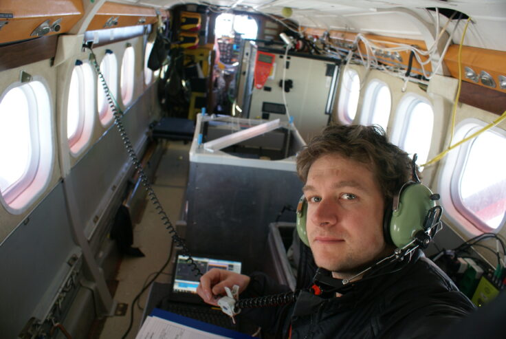 Tom Jordan takes a selfie inside a small plane. He is wearing comms headphones. The inside of the plane is very bare, other than instrumentation and wires connecting to instruments outside