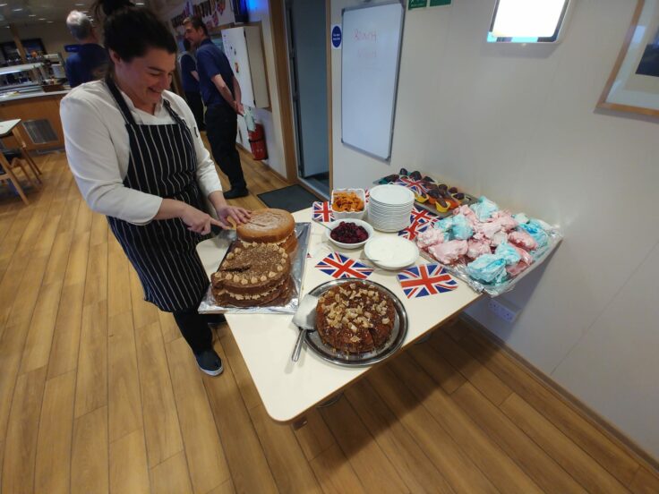 A woman wearing a striped apron stands next to a table covered in cakes. She is cutting a cake.