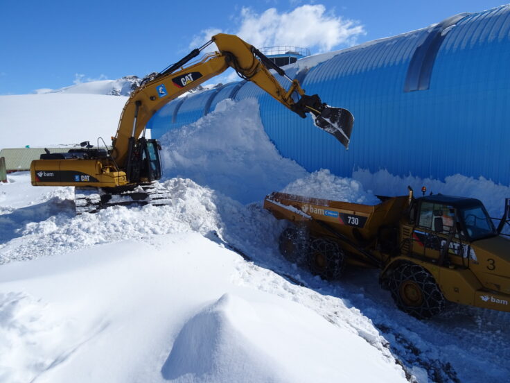 a yellow crane removing snow from the side of a blue building with a lot of snow piled up at the front