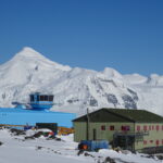 A blue building with a control tower on the roof and white snowy mountains in the background with a blue sky above