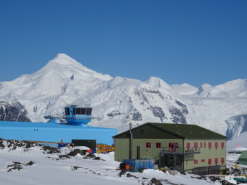 A blue building with a control tower on the roof and white snowy mountains in the background with a blue sky above