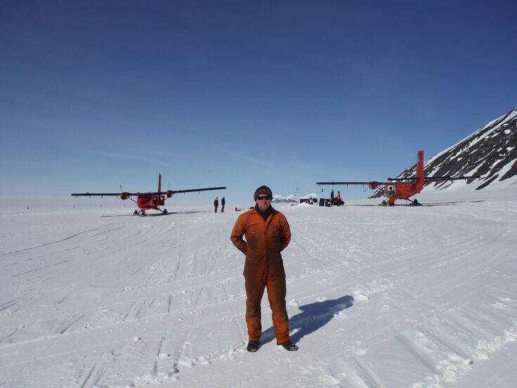 A man on a snow covered slope, with two Twin Otter planes behind him the distance