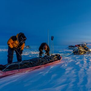 Two people attach gear to a trailer to be dragged by a skidoo on a frozen icey surface.