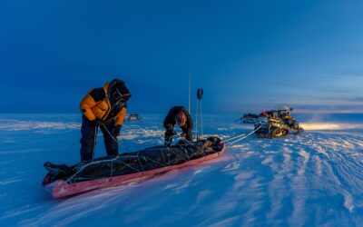 Two people attach gear to a trailer to be dragged by a skidoo on a frozen icey surface.