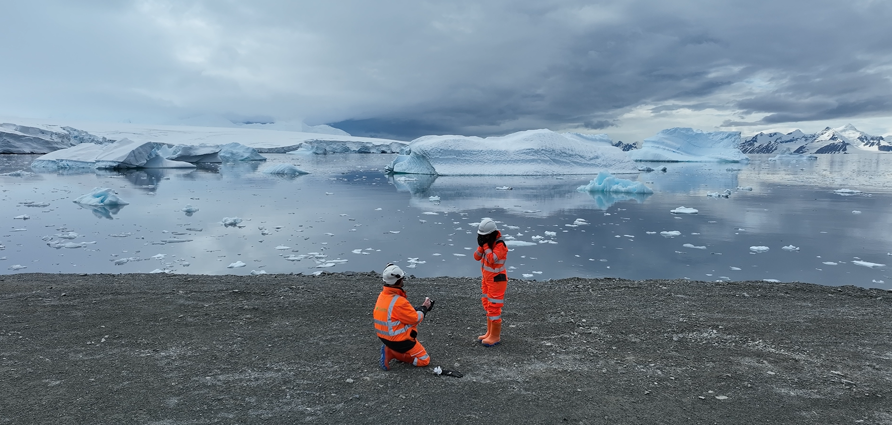 Two people standing on ground in front of an icy lake with snowy mountains behind.