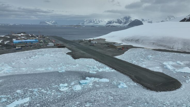 A short runway with snow covered ground to either side and snowy mountains and sea behind.
