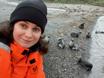 A smiling woman wearing a woolly hat on a rocky beach with some seals