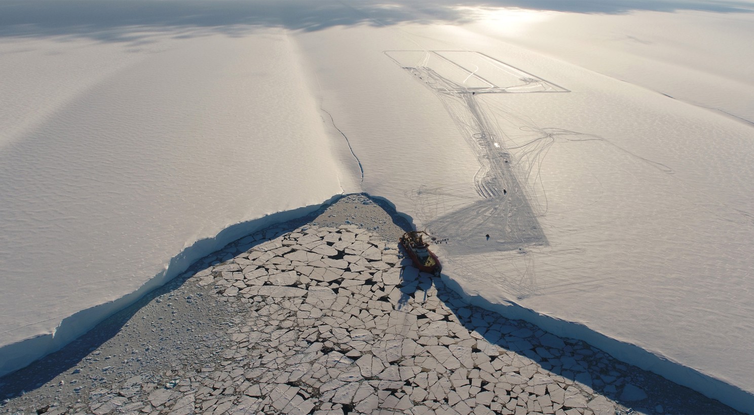 A drone image of a ship looking very small against an ice shelf. 