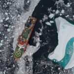 An aerial view of a red ship, sailing through dark water, covered with ice bergs in various sizes.