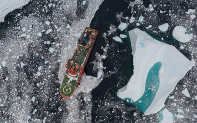 An aerial view of a red ship, sailing through dark water, covered with ice bergs in various sizes.