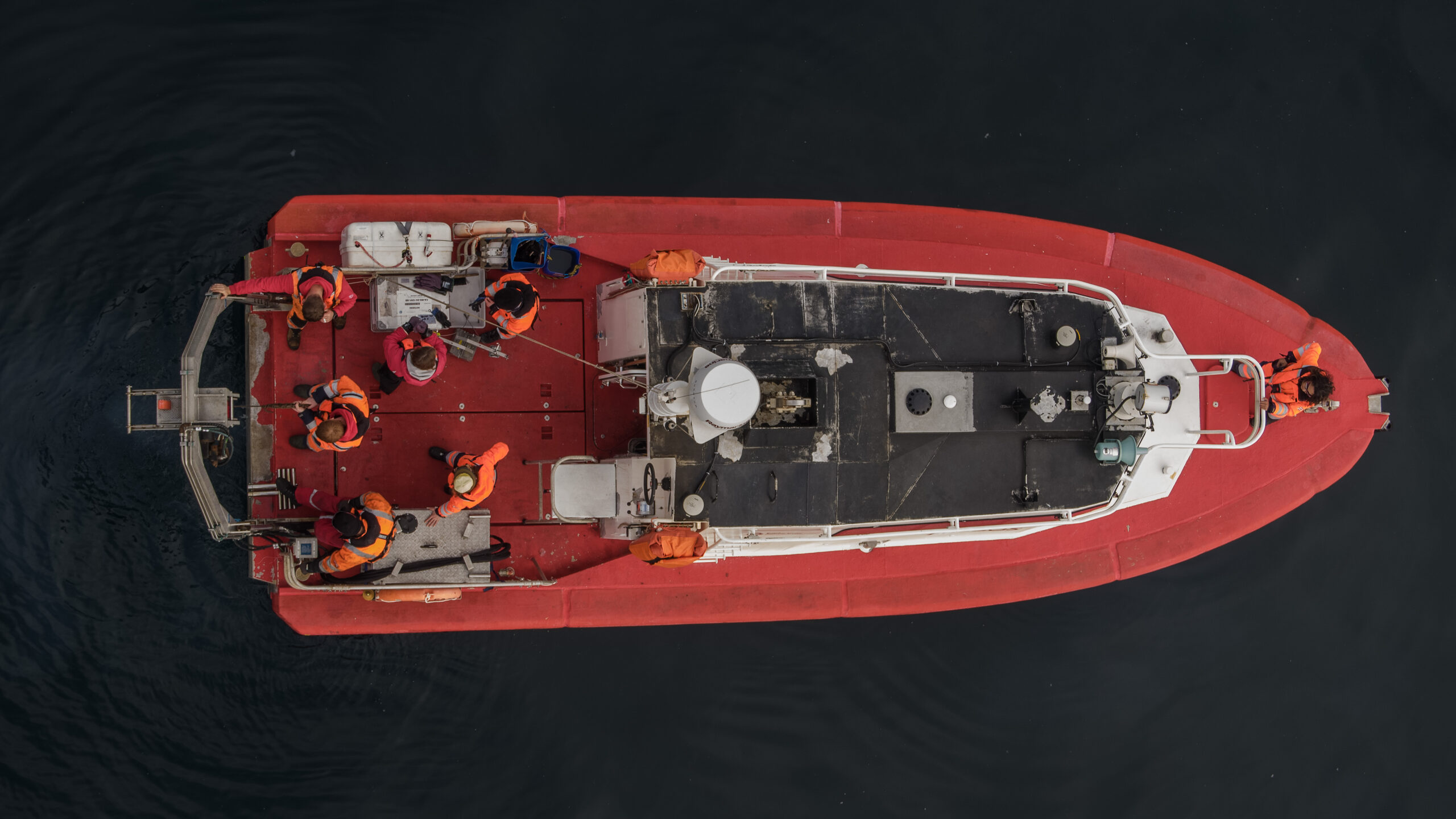 An aerial shot into a small red boat with people sitting inside, on very calm dark water.