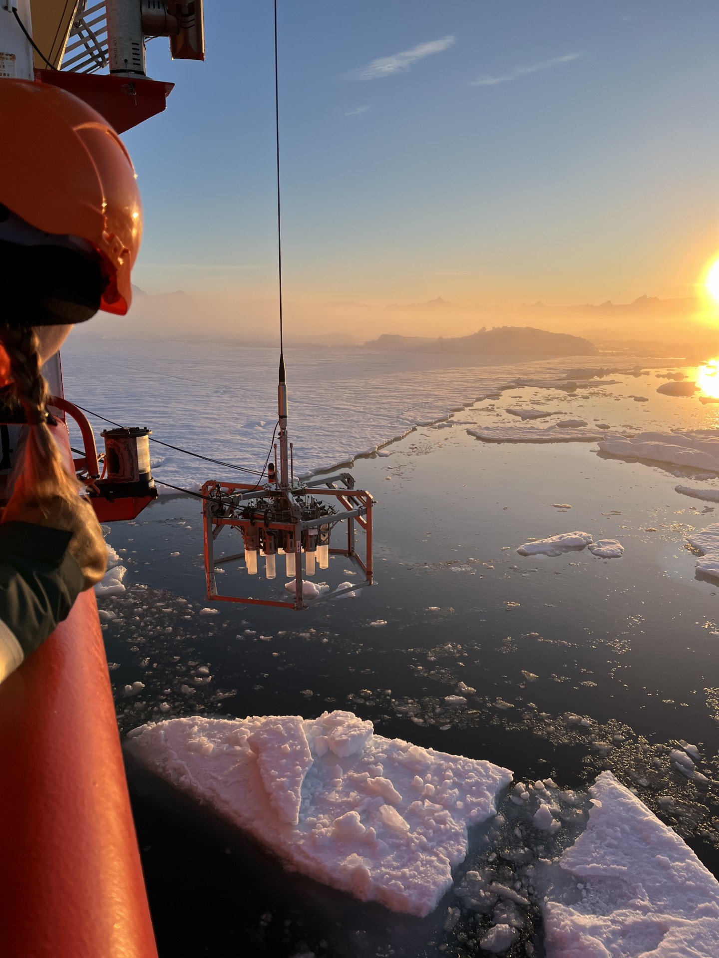 A metal sampling instrument is hanging beside a boat, above a body of water with ice on the surface, a sunset in the background.