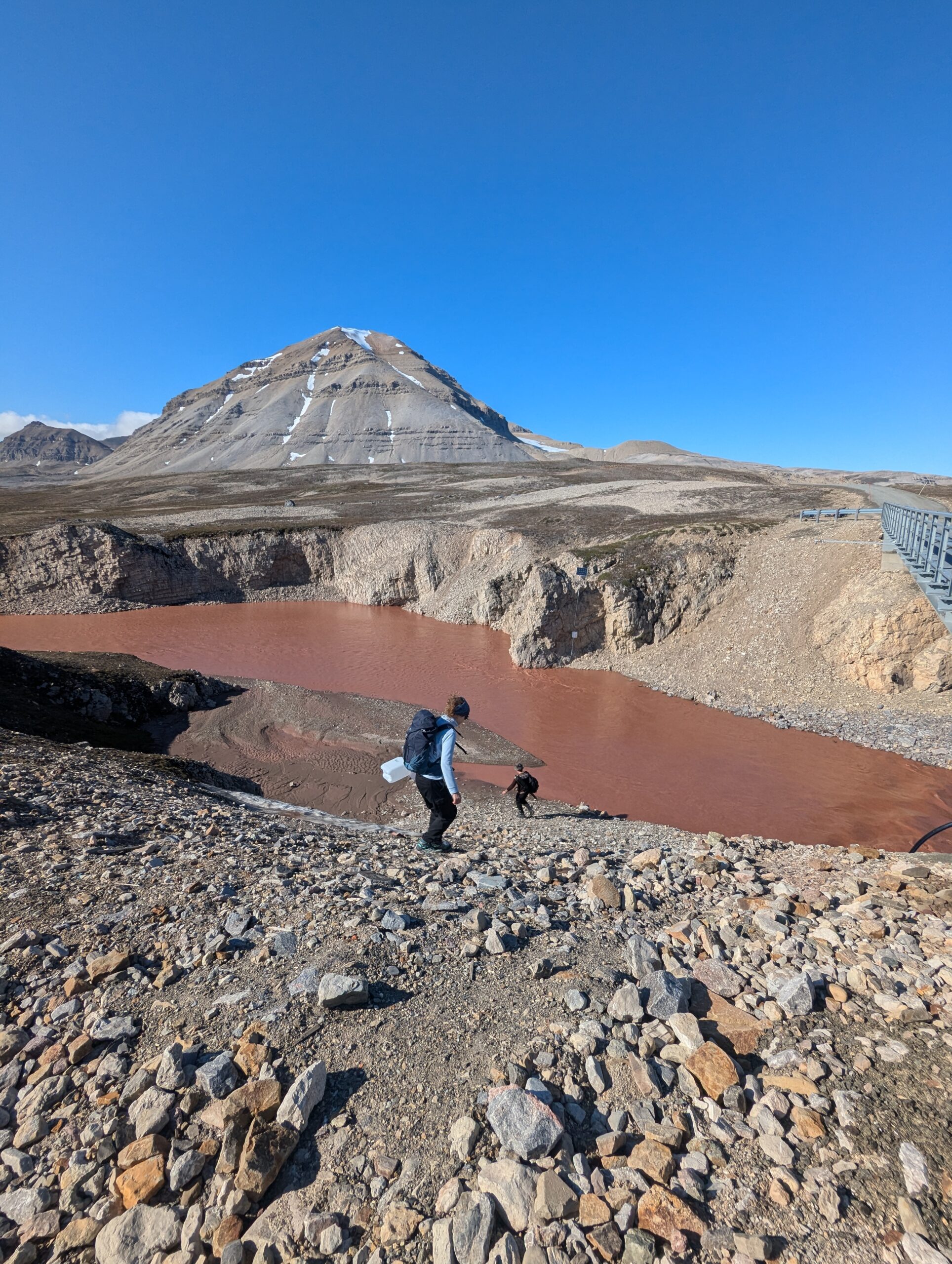 A muddy river flows through a rocky valley, two people descend the valley.