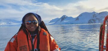 A person posing for the camera and standing in front of a body of water with ice covered mountains behind.