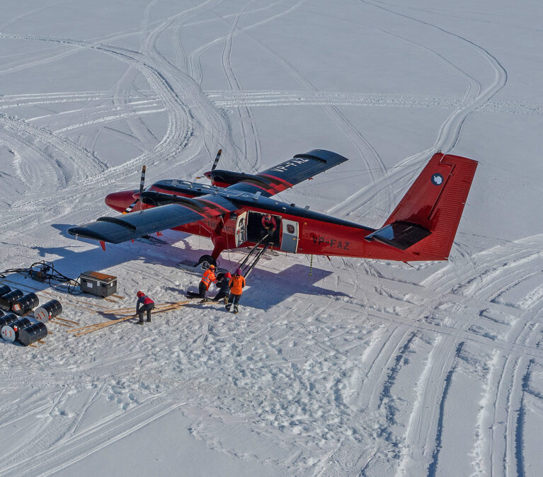 A red Twin Otter aircraft loading cargo including fuel drums at Gromits Creek, Antarctica.
